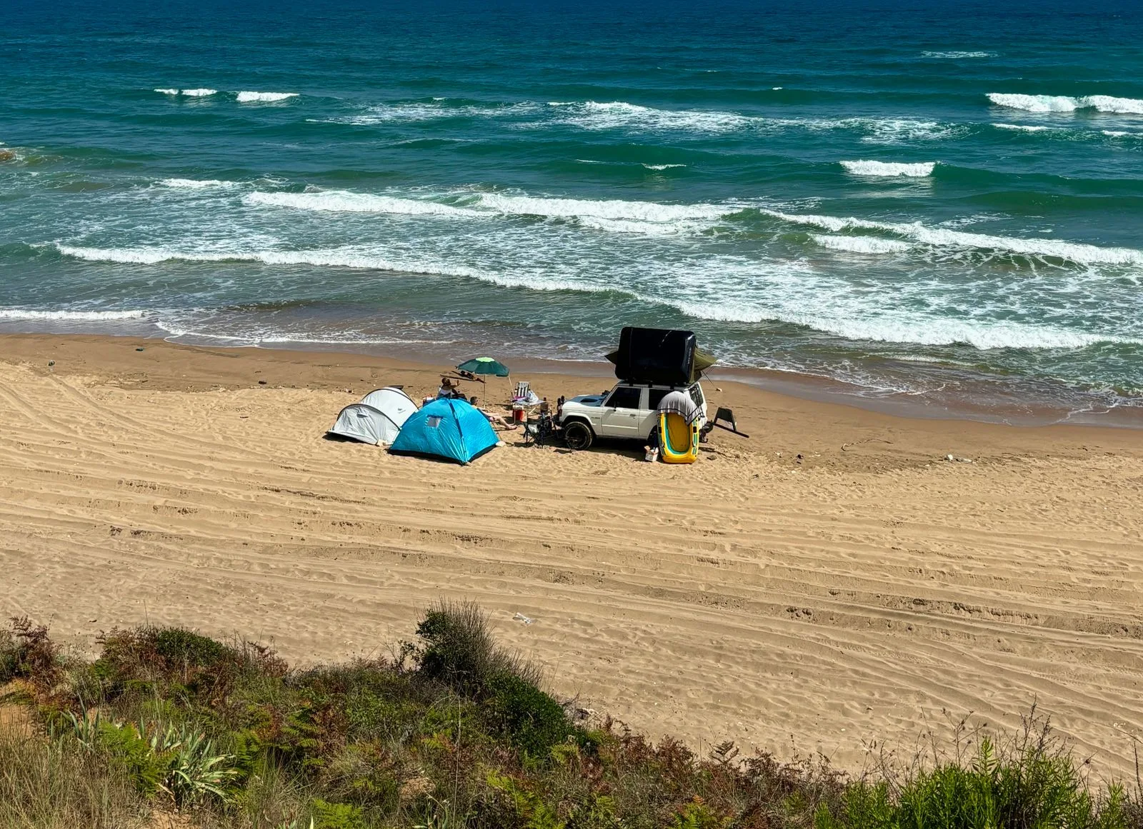 Tentes et un véhicule installés sur une plage de sable au bord d'une mer turquoise, en été