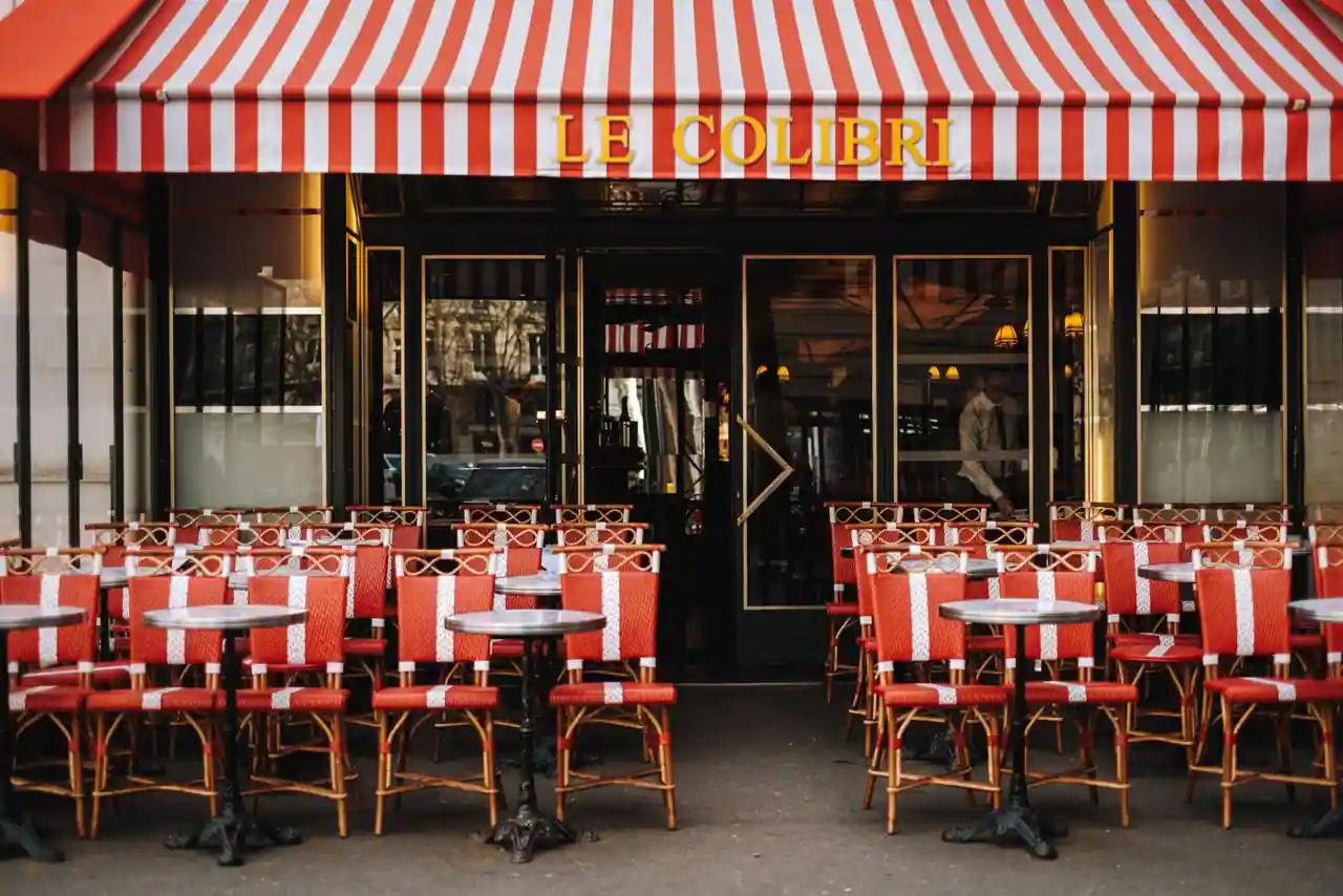 Terrasse d'un café parisien sous son store rayé, avec chaises en rotin et petites tables rondes sur le trottoir.