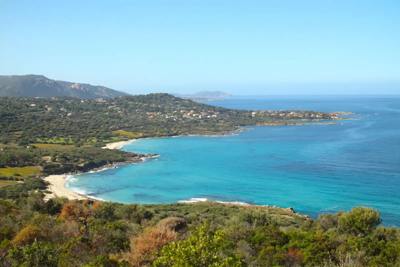 Littoral corse couvert de maquis, avec une crique de sable clair et une mer turquoise en contrebas