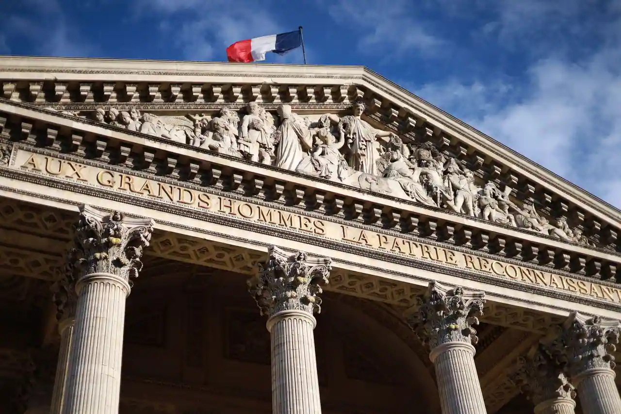 Drapeau tricolore français déployé au sommet du fronton du Panthéon, à Paris, sous un ciel bleu.