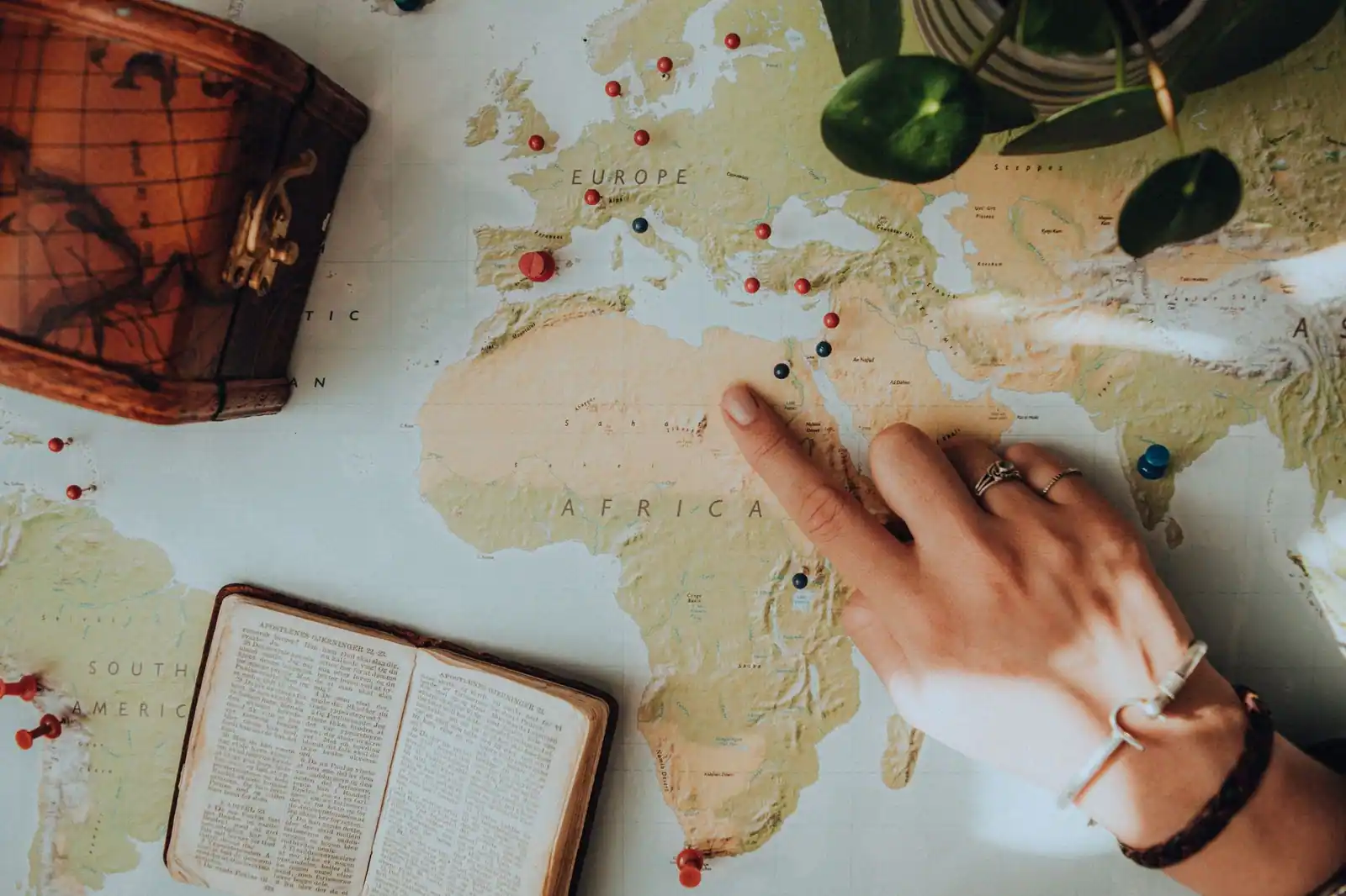 Main désignant un point sur une carte du monde constellée d'épingles de couleur, près d'un livre ancien ouvert.