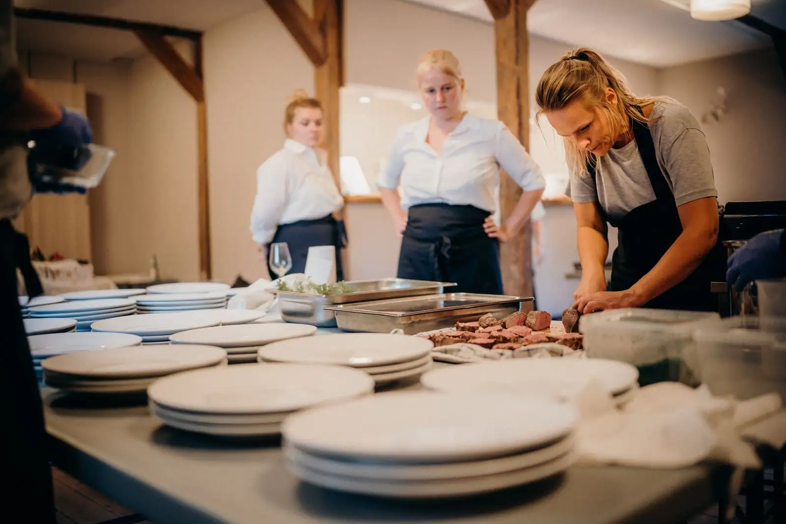 Cuisine professionnelle pendant un service : piles d'assiettes au passe, viande tranchée, personnel en tablier.