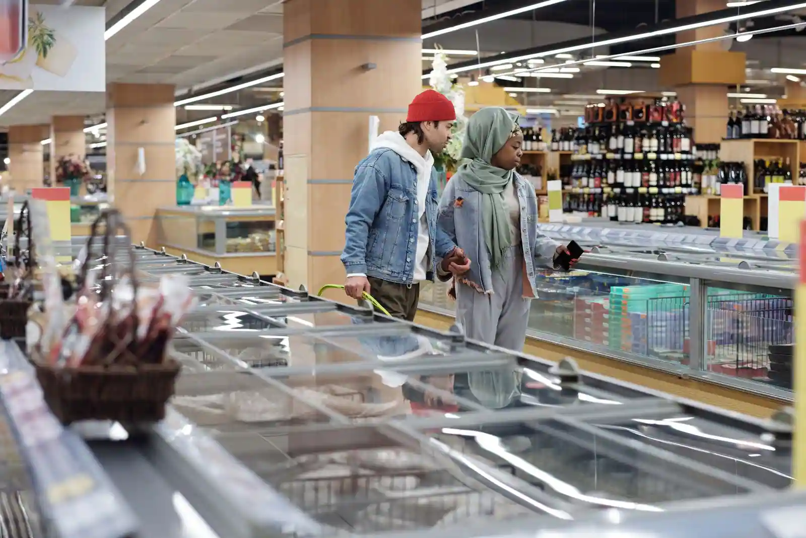 Deux clients longent les bacs à surgelés vitrés d'un magasin alimentaire, un panier de courses à la main