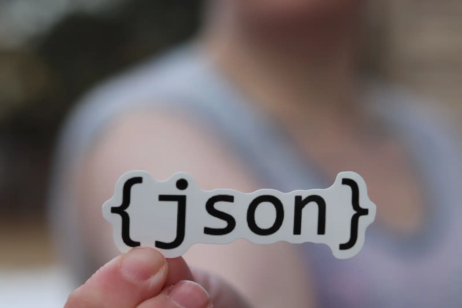 Autocollant portant le mot json entre accolades, tenu entre les doigts devant un fond flou