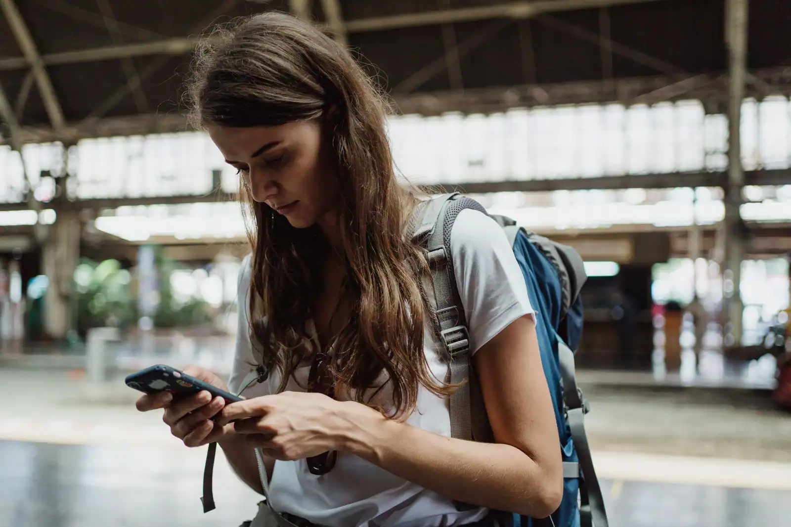 Une voyageuse avec un sac à dos consulte l'écran de son téléphone sous la verrière d'une gare.